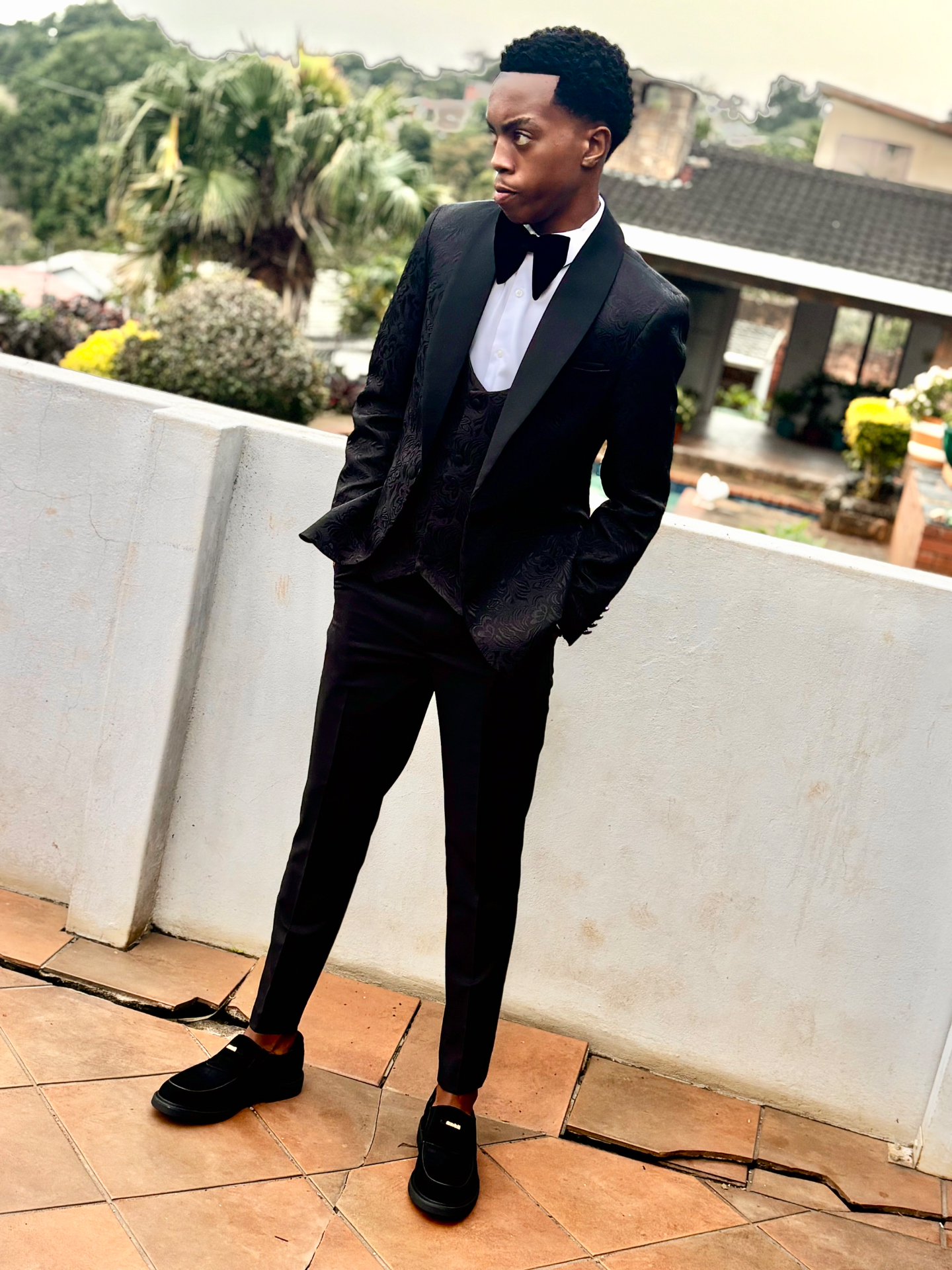 Sipho Biyela in a patterned black tuxedo leaning against a wall outdoors